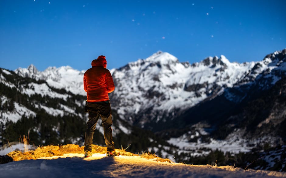 A lone hiker in a red jacket stands illuminated, gazing at the majestic snowy mountains under a starry night sky.